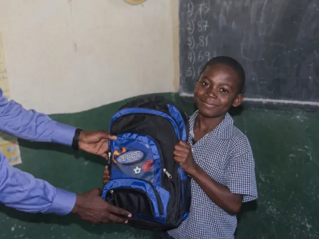 Un niño con camisa a cuadros recibe una mochila nueva en su salón de clases